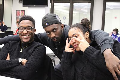 三个非裔美国学生摆姿势拍照. 左边的一名女学生正在用两只手做v形手势，并把它们放在眼睛附近. 中间的一名男学生弯下腰，双臂搂着另外两名学生. 右边的学生双臂交叉，笑容灿烂. 